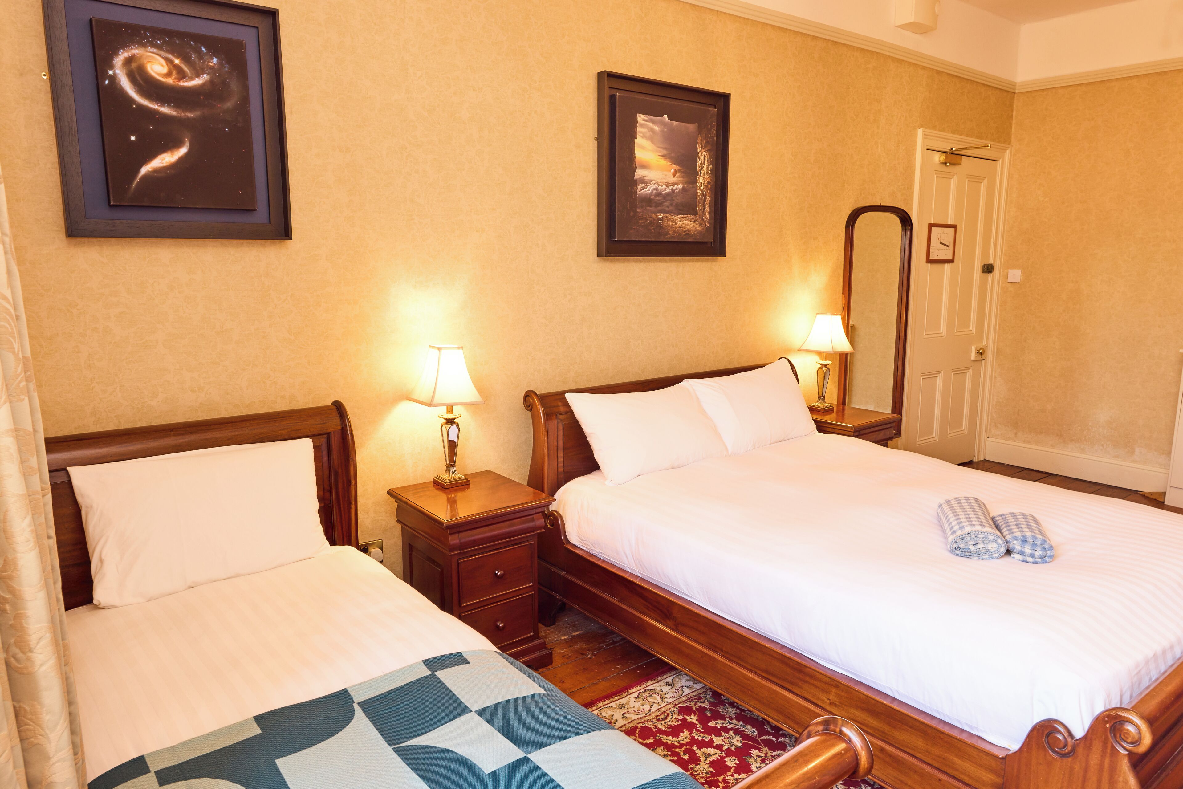 A hotel room with two beds, a wooden headboard, a nightstand with a lamp, a framed picture on the wall, and a door with a mirror.