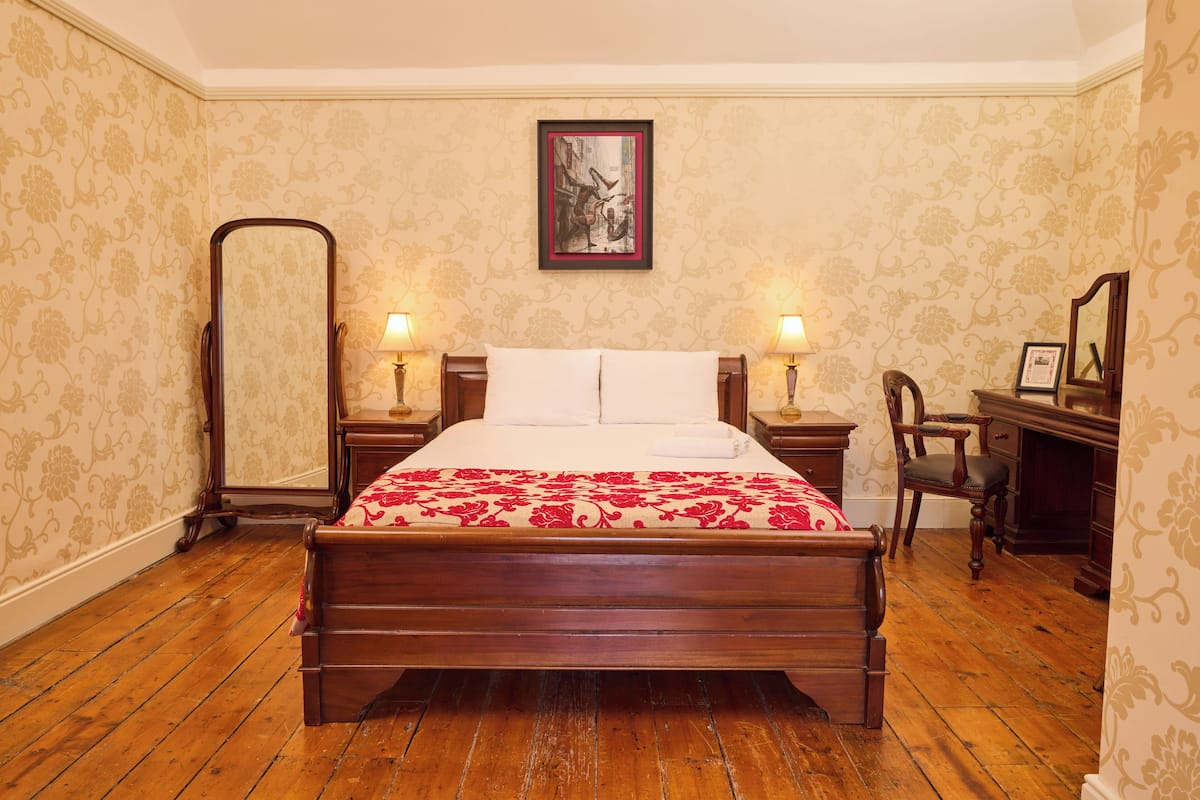 Una cama de madera clásica con colcha estampada, flanqueada por mesitas de noche con lámparas, en una habitación con papel tapiz estampado y suelo de madera.