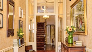 Un couloir avec une moquette rouge, une console en bois ornée de livres et d’un vase de fleurs, un miroir et un escalier menant à une autre pièce.