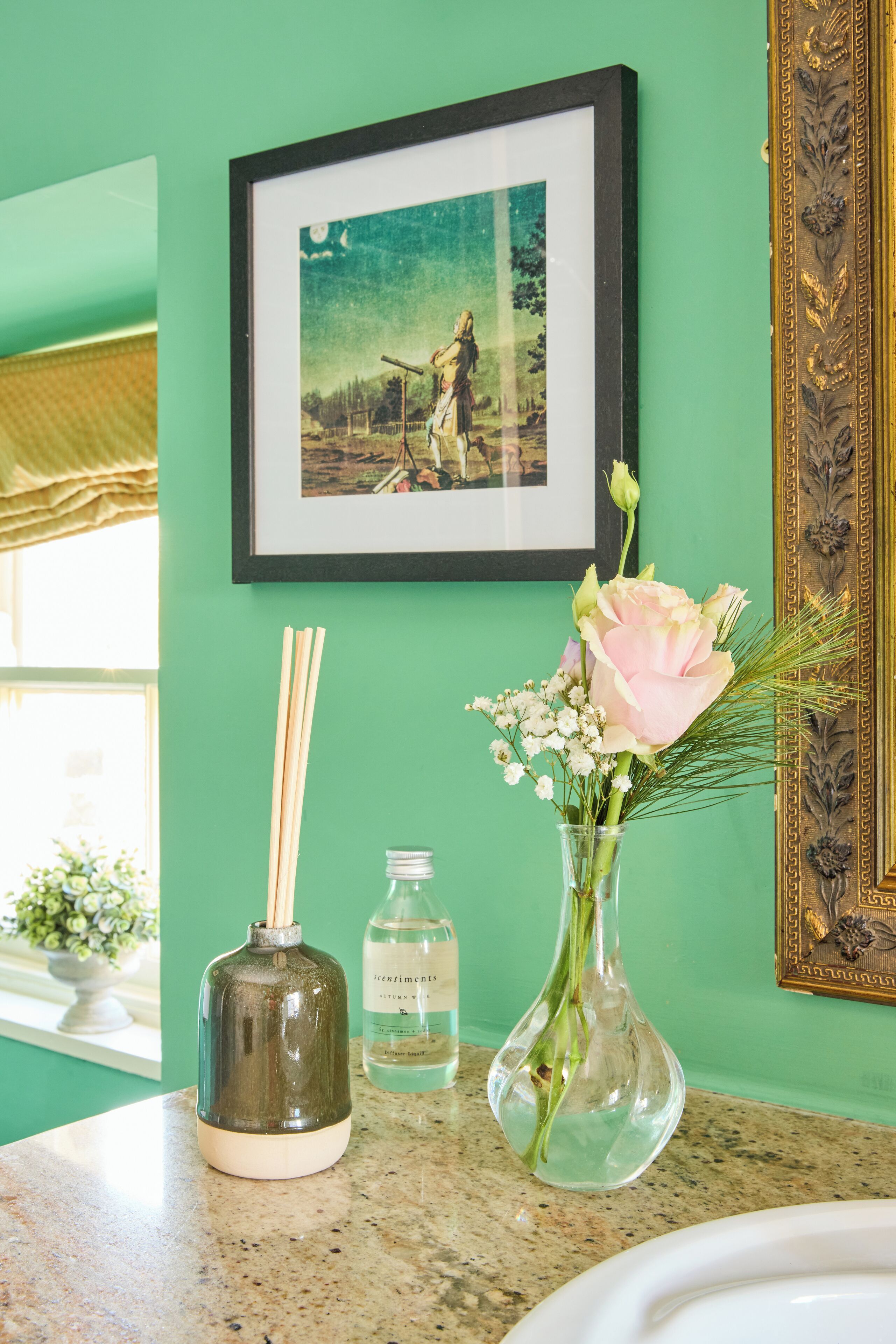 A bathroom with a green wall, a framed picture, a vase with flowers, a bottle, and a diffuser.