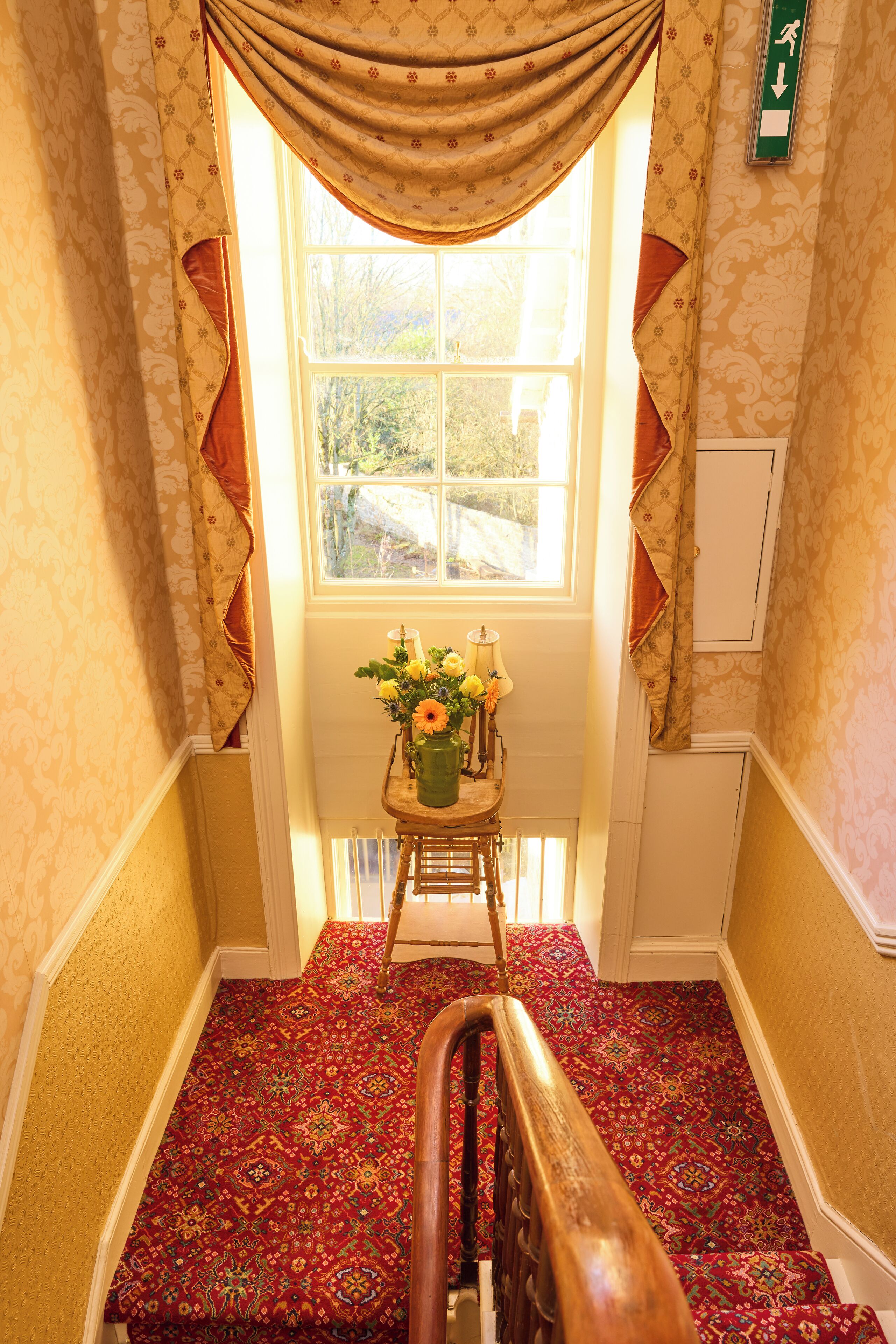 A carpeted staircase with a wooden handrail, a small table with flowers, and a window with curtains.