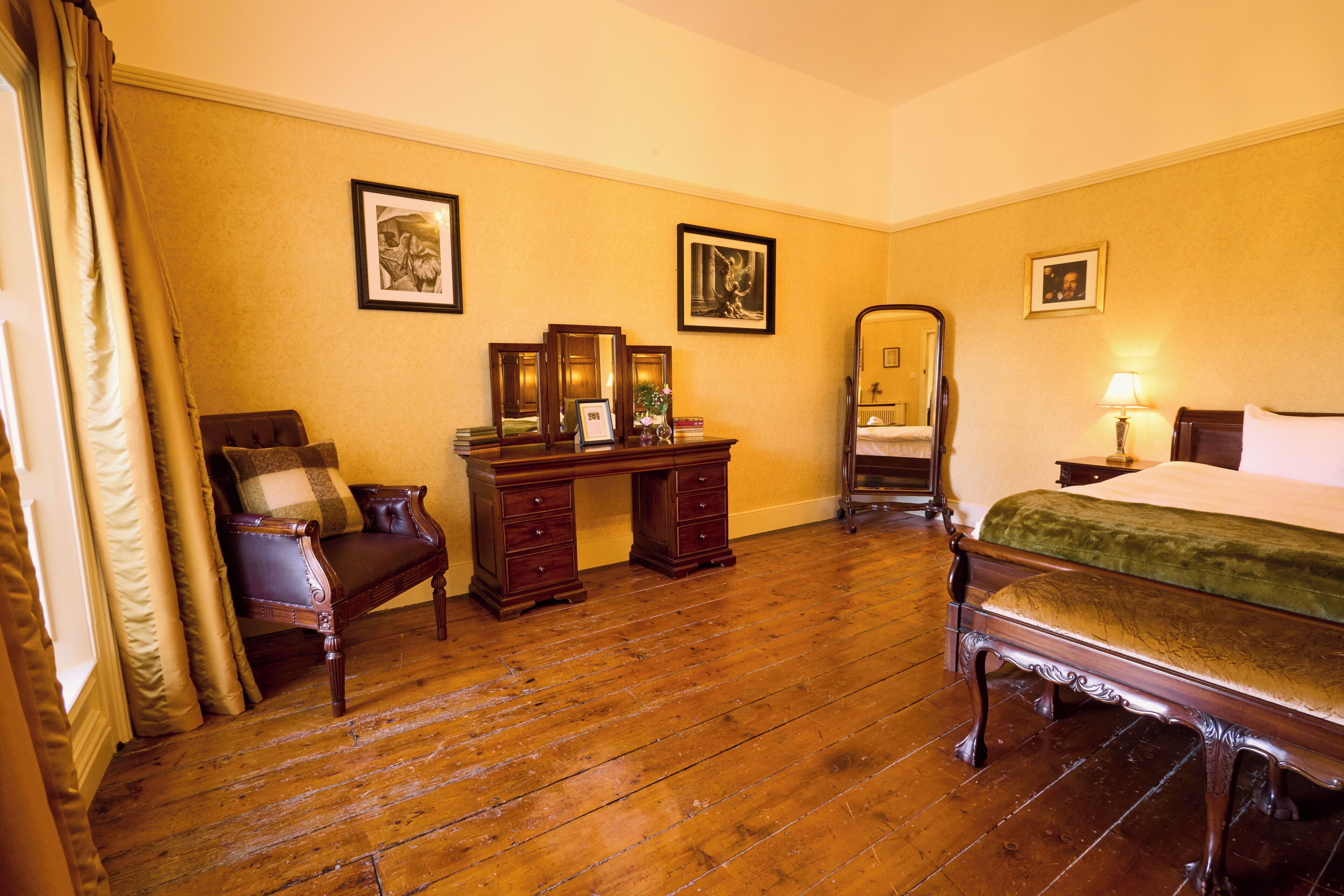 A traditional room with a wooden floor, a bed with a green bedspread, a desk with drawers, a chair, and a mirror.