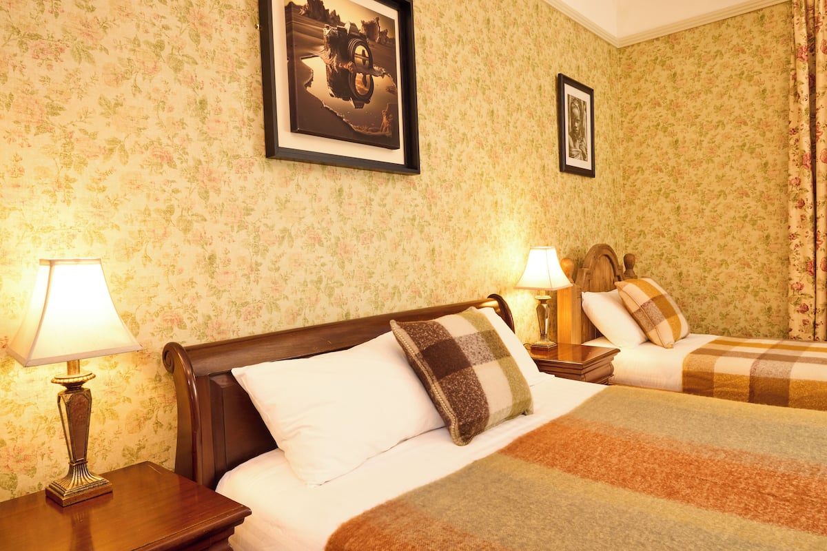Habitación de hotel con papel tapiz floral, dos camas con colchas a cuadros y cuadros enmarcados en las paredes.
