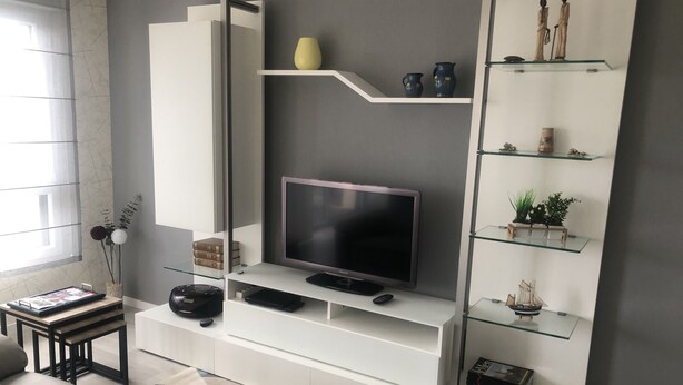 A modern living room with a flat-screen TV, white shelving units, and decorative items. - Magnifique F2 vue Port L'ostrea (Courseulles-sur-Mer)