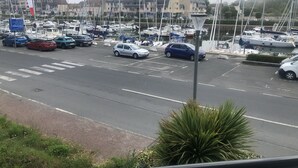 A marina with parked cars and boats, a pedestrian crossing, and buildings in the background. - Magnifique F2 vue Port L'ostrea (Courseulles-sur-Mer)