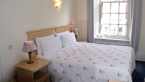A hotel room with a bed, bedside lamps, a nightstand, a window with a view of buildings, and a wall-mounted light. - Luxury Serviced Apartment with Daily Room Clean, Staffed Reception, King Size Be (London)