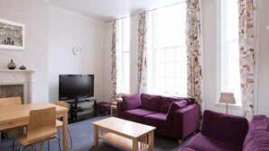Ein Wohnzimmer mit Kamin, Fernseher, Esstisch mit Stühlen und einem violetten Sofa.