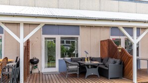 An outdoor patio with a wooden deck, a grill, wicker furniture, and a view of the surrounding greenery. - Beautiful apartment in Nexø (Nexø)