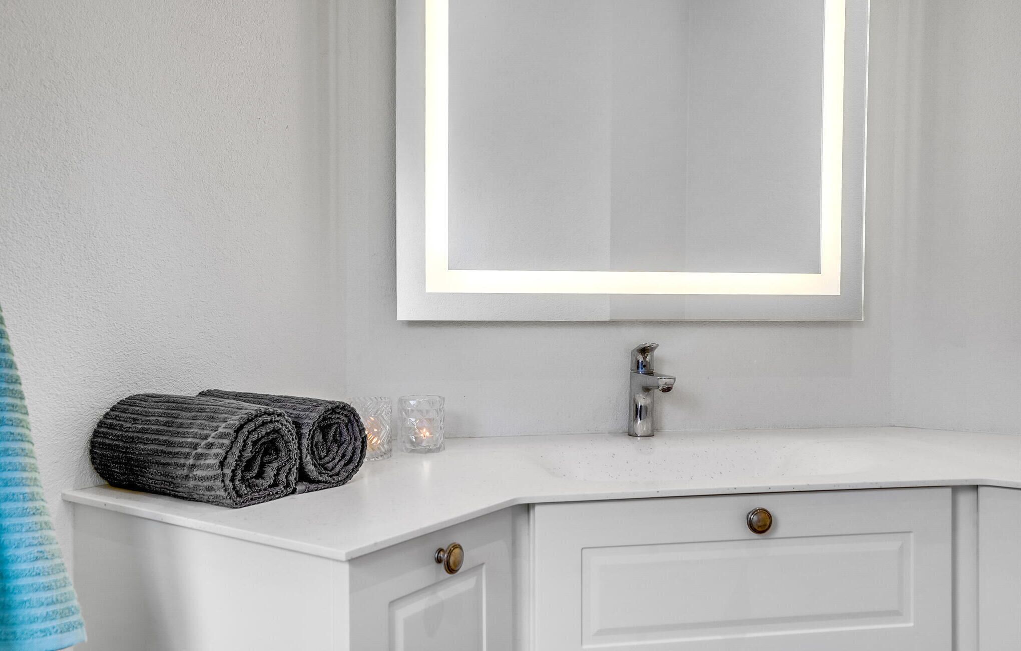Ein Badezimmer mit weißem Waschtisch, einem Spiegel mit Beleuchtung und einem Handtuch auf der Ablagefläche.