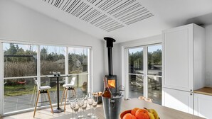 A modern kitchen with a wood-burning stove, large windows, and a dining area with chairs and wine glasses. - Awesome home in Blåvand with sauna (Blåvand)