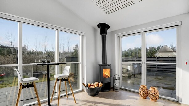 A modern room with large windows, a black wood-burning stove, a table with chairs, and a view of the outdoors. - Awesome home in Blåvand with sauna (Blåvand)