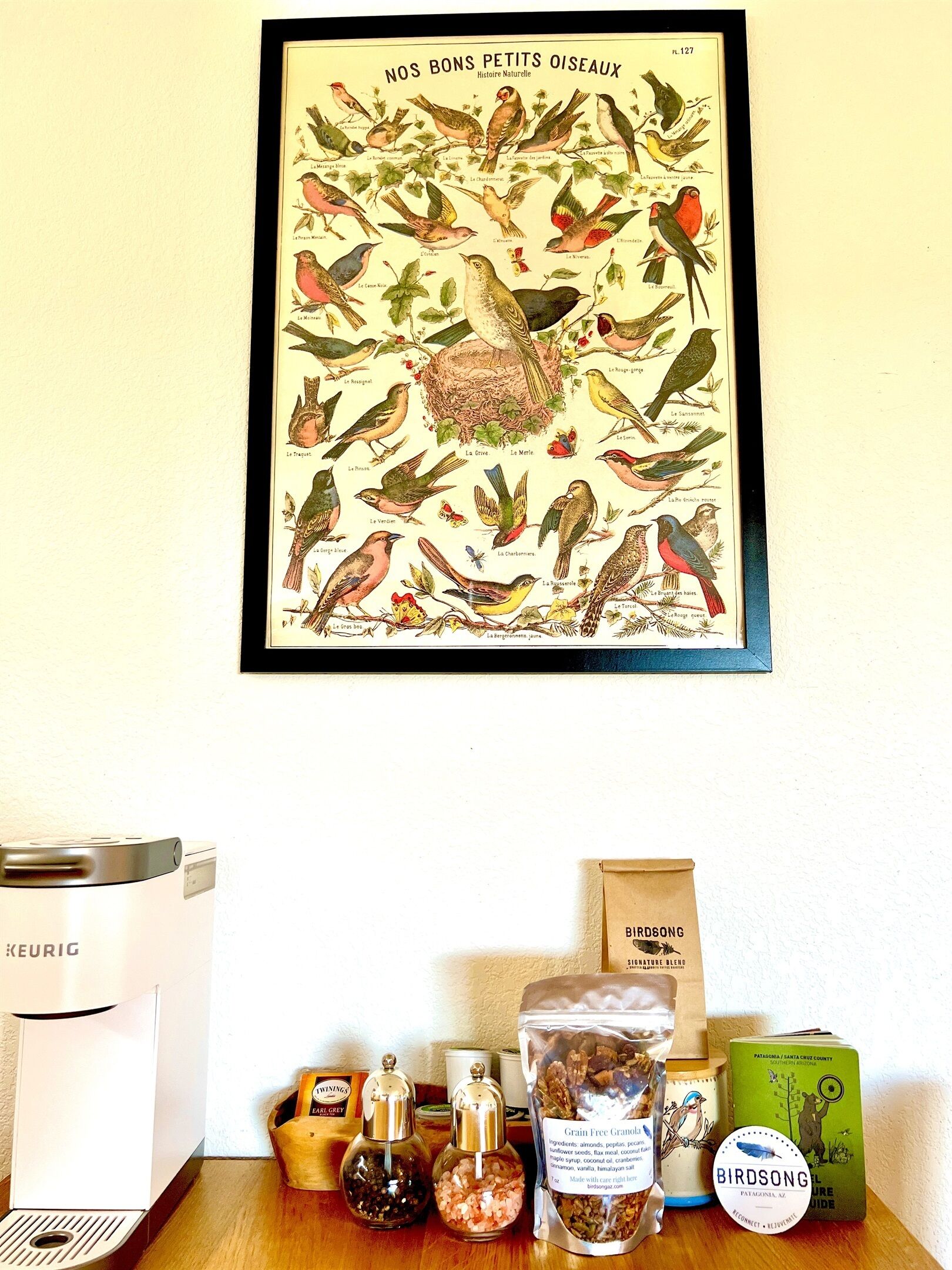 Ein gerahmtes Vogelbild an der Wand, eine Keurig-Kaffeemaschine und verschiedene Speisen auf einem Holztisch.
