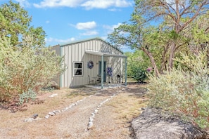 Une cabane rustique dotée d’un porche, entourée d’herbes sèches et d’arbres.