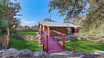 A wooden cabin with a red bridge, surrounded by a grassy area and trees.