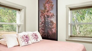Une chambre à coucher avec un lit, deux oreillers ornés de motifs floraux, une grande fenêtre avec des stores et une affiche représentant un arbre aux fleurs roses.