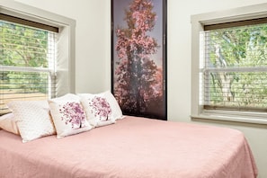 A bedroom with a bed, two pillows with floral designs, a large window with blinds, and a poster of a tree with pink flowers. - Lavender - Private Getaway with Horseshoes, Stargazing, and Walking Trails (Wimberley)