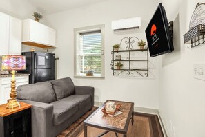 A compact living space with a gray sofa, a small coffee table, a decorative lamp, a wall-mounted TV, and a window with blinds.