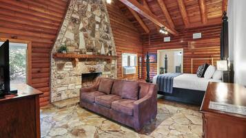 A log cabin interior with a stone fireplace, a sofa, a bed, and a wooden ceiling.