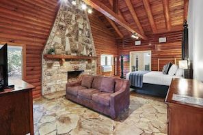 Ein Blockhaus-Interieur mit Kamin aus Stein, einer Couch, einem Bett und einer Holzbalkendecke.