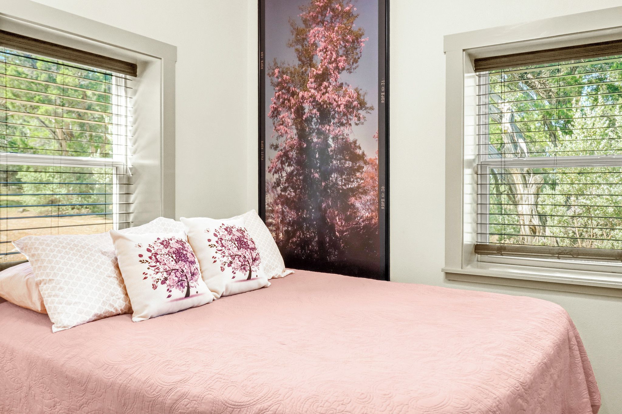 Een slaapkamer met een bed, twee kussens met bloemenmotieven, een groot raam met jaloezieën en een poster van een boom met roze bloemen.