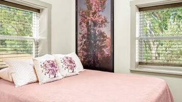 Een slaapkamer met een bed, twee kussens met bloemenmotieven, een groot raam met jaloezieën en een poster van een boom met roze bloemen.