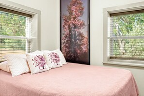 Une chambre à coucher avec un lit, deux oreillers ornés de motifs floraux, une grande fenêtre avec des stores et une affiche représentant un arbre aux fleurs roses.