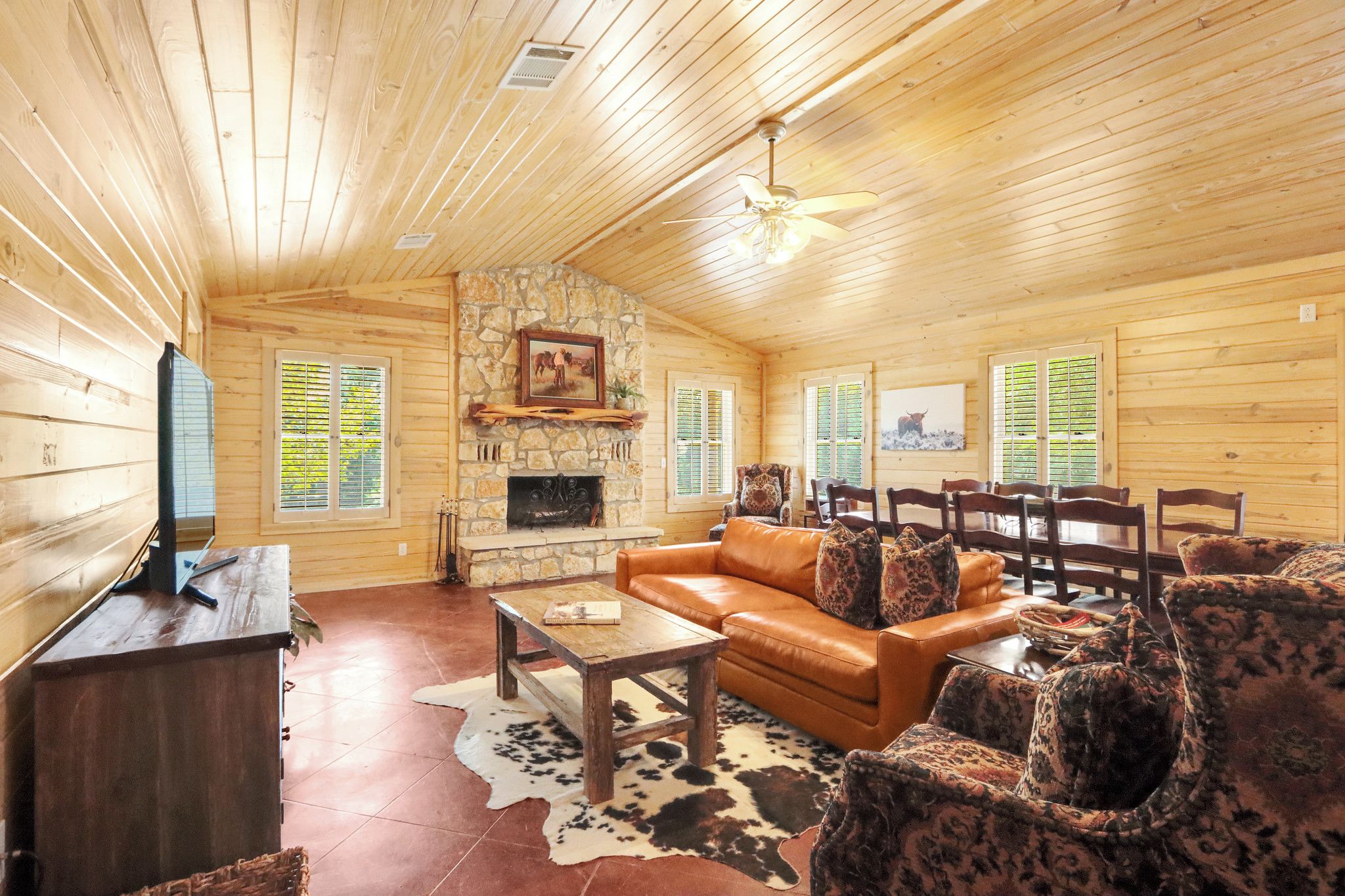 A cozy log cabin interior with a leather sofa, patterned chairs, a stone fireplace, and wooden walls.