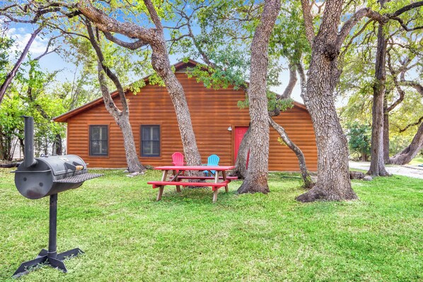 A wooden cabin with a picnic table and chairs, surrounded by trees. - The Oak Lodge - 3 Bedroom Cabin with Swimming Pool Access! Close to Downtown (Wimberley)