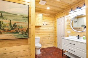A wooden cabin bathroom with a white toilet, a sink with a mirror, and a painting of a desert landscape. - The Oak Lodge - 3 Bedroom Cabin with Swimming Pool Access! Close to Downtown (Wimberley)