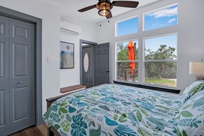 A bedroom with a bed, a ceiling fan, a window with a view of the outdoors, and a door.