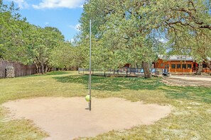 A fenced outdoor area with a sandpit, trees, and a building in the background. - The Oak Lodge - 3 Bedroom Cabin with Swimming Pool Access! Close to Downtown (Wimberley)