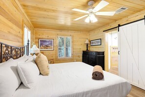A wooden cabin room with a bed, a ceiling fan, a window with shutters, and a door.