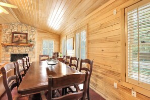 A wooden-paneled dining room with a stone fireplace, wooden ceiling, and large windows with shutters.