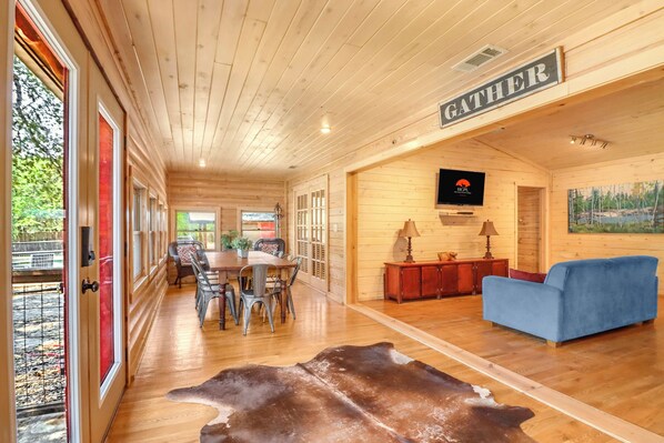 A spacious room with a wooden ceiling, a dining area with chairs and a table, a blue sofa, a mounted television, and a sign that reads 'GATHERER'.