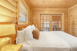 Un dormitorio de cabaña de madera con una cama, almohadas, una lámpara y una ventana con persianas.