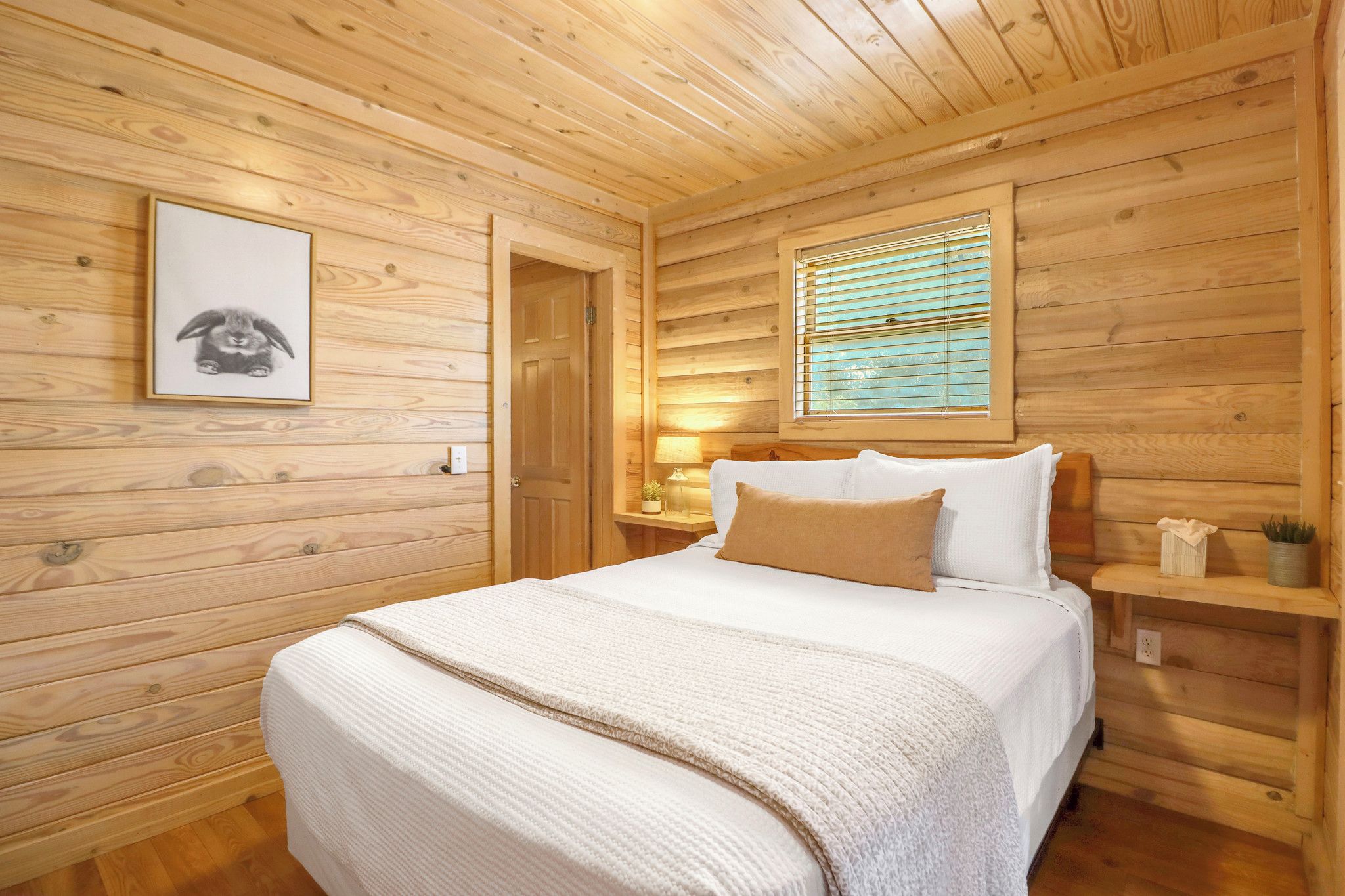 Een slaapkamer in een houten blokhut met een bed, een lijst met een foto aan de muur, een raam met zonwering en een nachtkastje met een lamp.