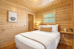 Ein Schlafzimmer einer Holzhütte mit einem Bett, einem gerahmten Bild an der Wand, einem Fenster mit Jalousien und einem Nachttisch mit einer Lampe.