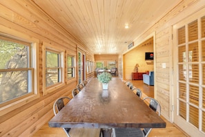 Une longue table  à manger en bois, avec des chaises, une plante en pot et un téléviseur fixé au mur.