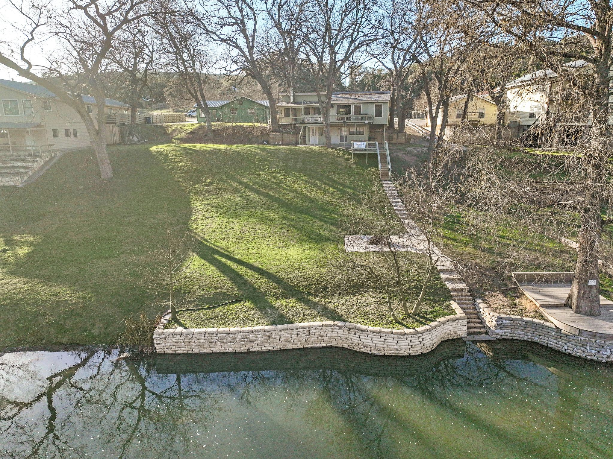 Un quartier résidentiel caractérisé par une pente herbeuse, un mur de soutènement en pierre et un petit plan d’eau où se reflètent les arbres.