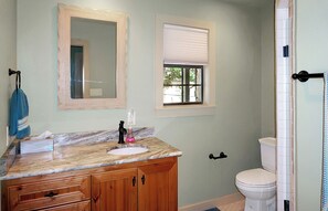 A bathroom with a marble sink, wooden cabinets, a toilet, a window with blinds, and a shower curtain. - Lazy River - Relaxing Accommodations for 6 on Blanco River (Wimberley)