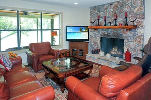 Un salón con mobiliario de cuero, chimenea de piedra, televisor y un ventanal con vistas al exterior.