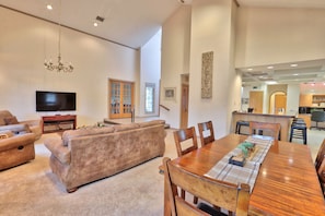 A spacious living room with a large sofa, a dining table with chairs, a chandelier, and a flat-screen TV.