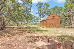 A wooden barn with a sloped roof, surrounded by trees and a dirt path. - Lavender - Private Getaway with Horseshoes, Stargazing, and Walking Trails (Wimberley)