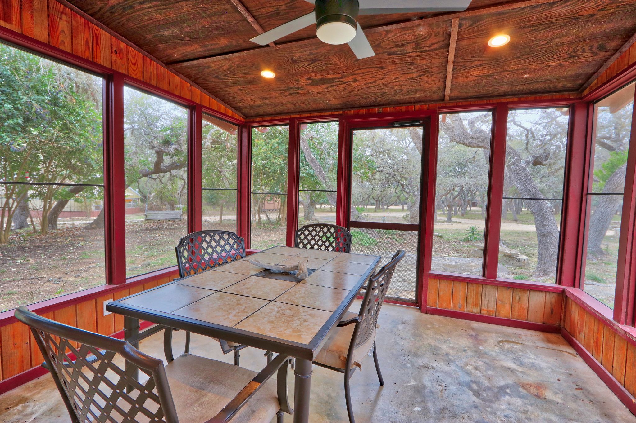 A covered patio with a wooden table and chairs, large windows, and a ceiling fan.