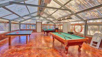 Amplia zona recreativa interior con varias mesas de billar y una mesa de ping pong, que cuenta con un techo alto con estructura metálica y grandes ventanales que permiten la entrada de luz natural.