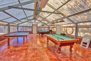 A spacious indoor recreational area with multiple pool tables and a ping pong table, featuring a high ceiling with a metal framework and large windows allowing natural light.