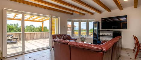 En rummelig opholdsstue med en lædersofa, et fjernsyn monteret på væggen og en balkon med udsigt over naturen.