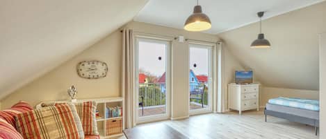 Ein Schlafzimmer im Loft-Stil mit schrägem Dach, einem Bett, einem Fernseher, einem Bücherregal, einer Uhr und einem Balkon mit Blick auf Häuser.