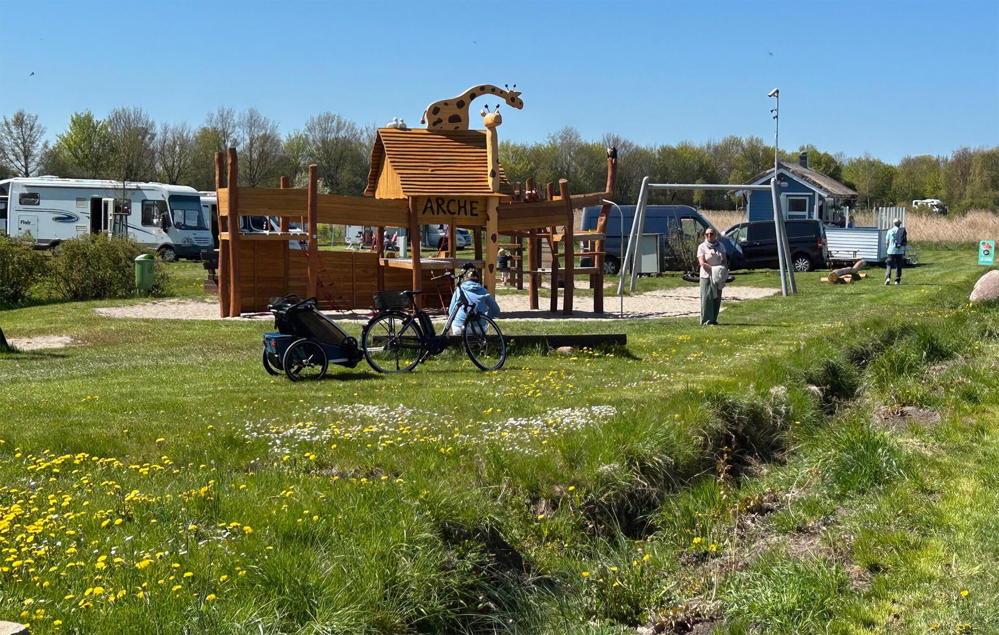 Een camping met een houten constructie waarop 'ARCHE' staat, fietsen en een aanhangwagen.