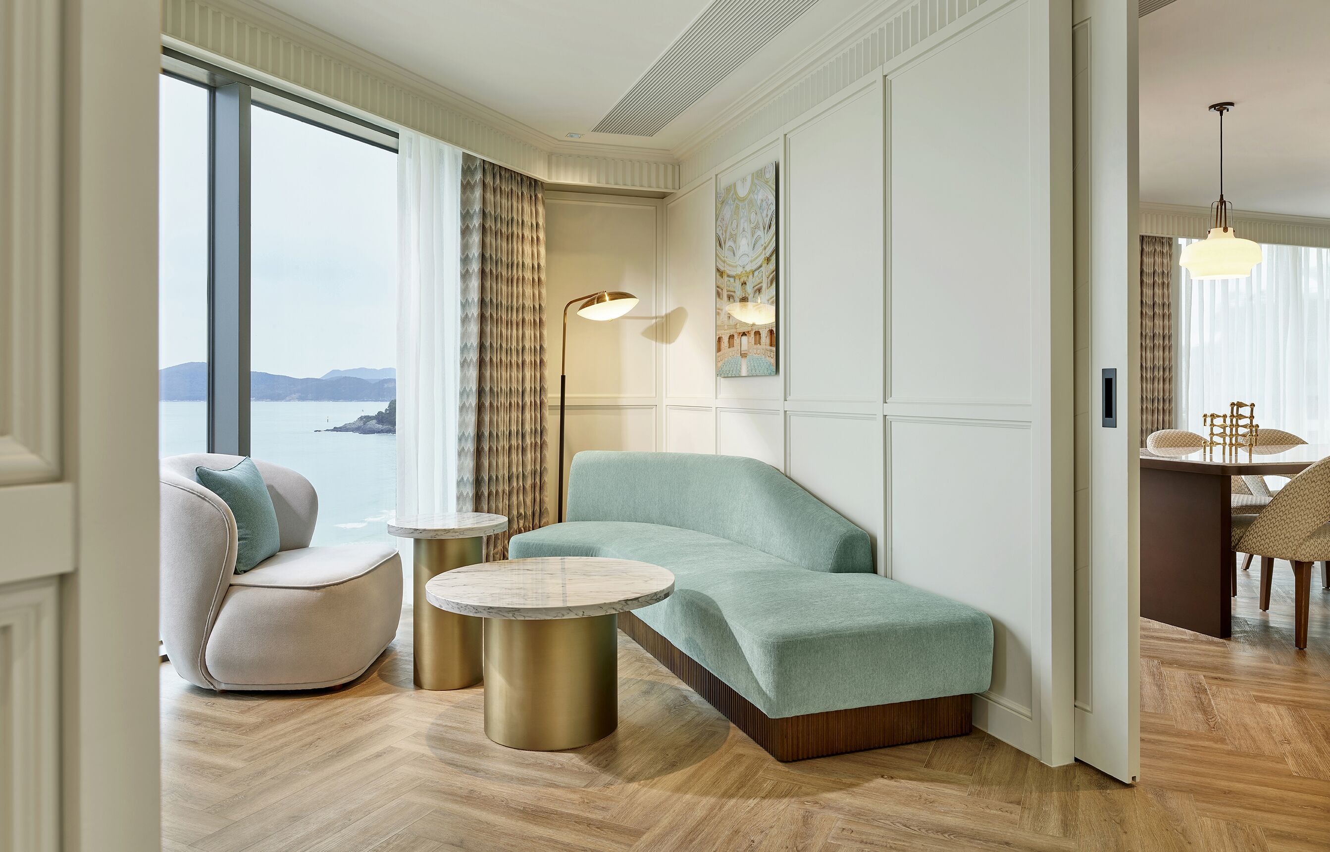 A modern hotel room with a large window, a sofa, a round coffee table, and a dining area with a chair.